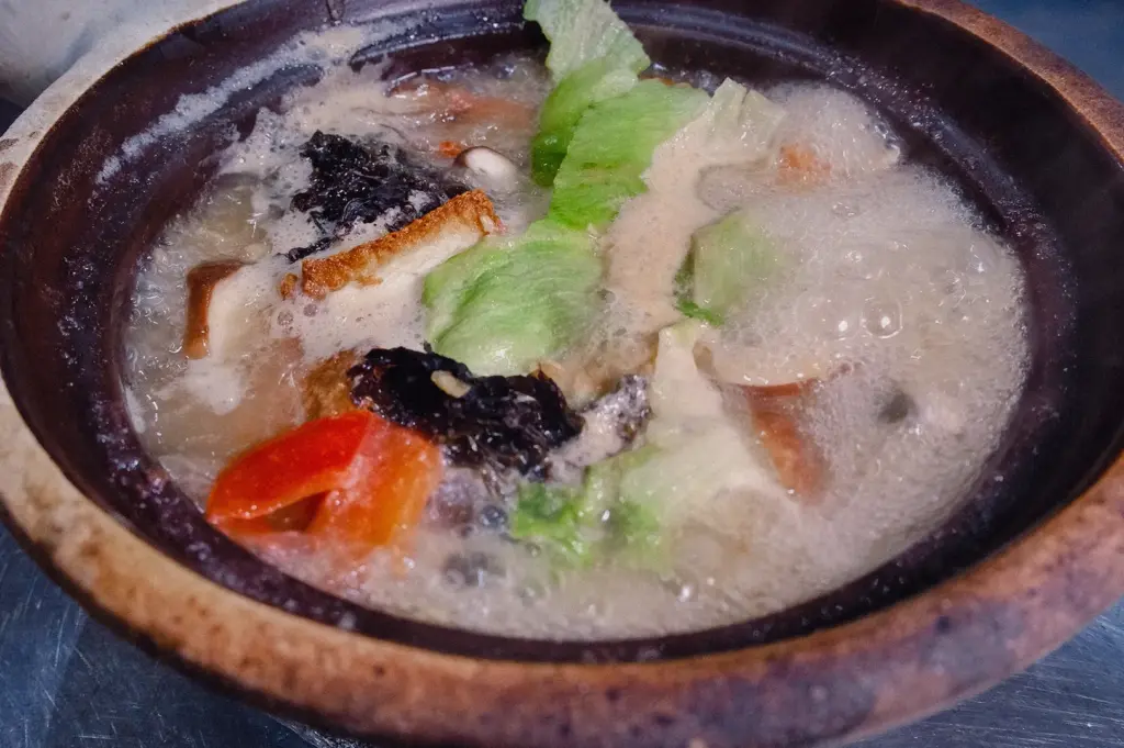 A high-angle close-up shot displays a freshly cooked claypot of Grandpa's Pomfret Soup bubbling gently with lettuce, tomatoes, and seaweed.